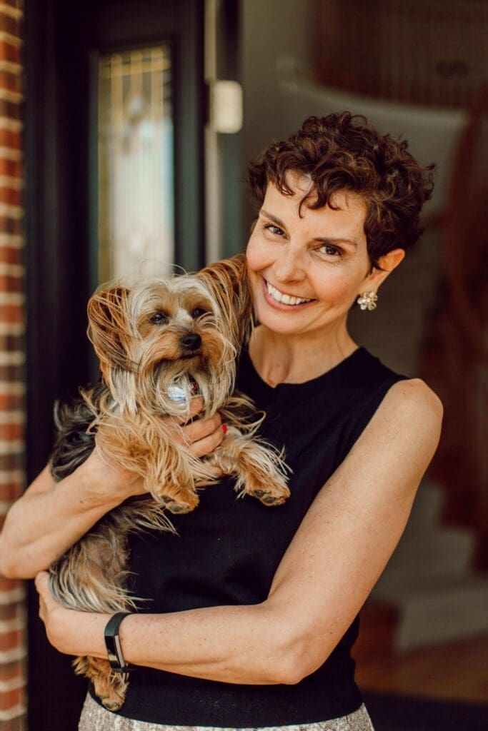 A woman with short curly hair smiles while holding a small, fluffy dog. Dressed in a black sleeveless top and earrings, she stands near an organized doorway, bricks peeking through the background indoors.