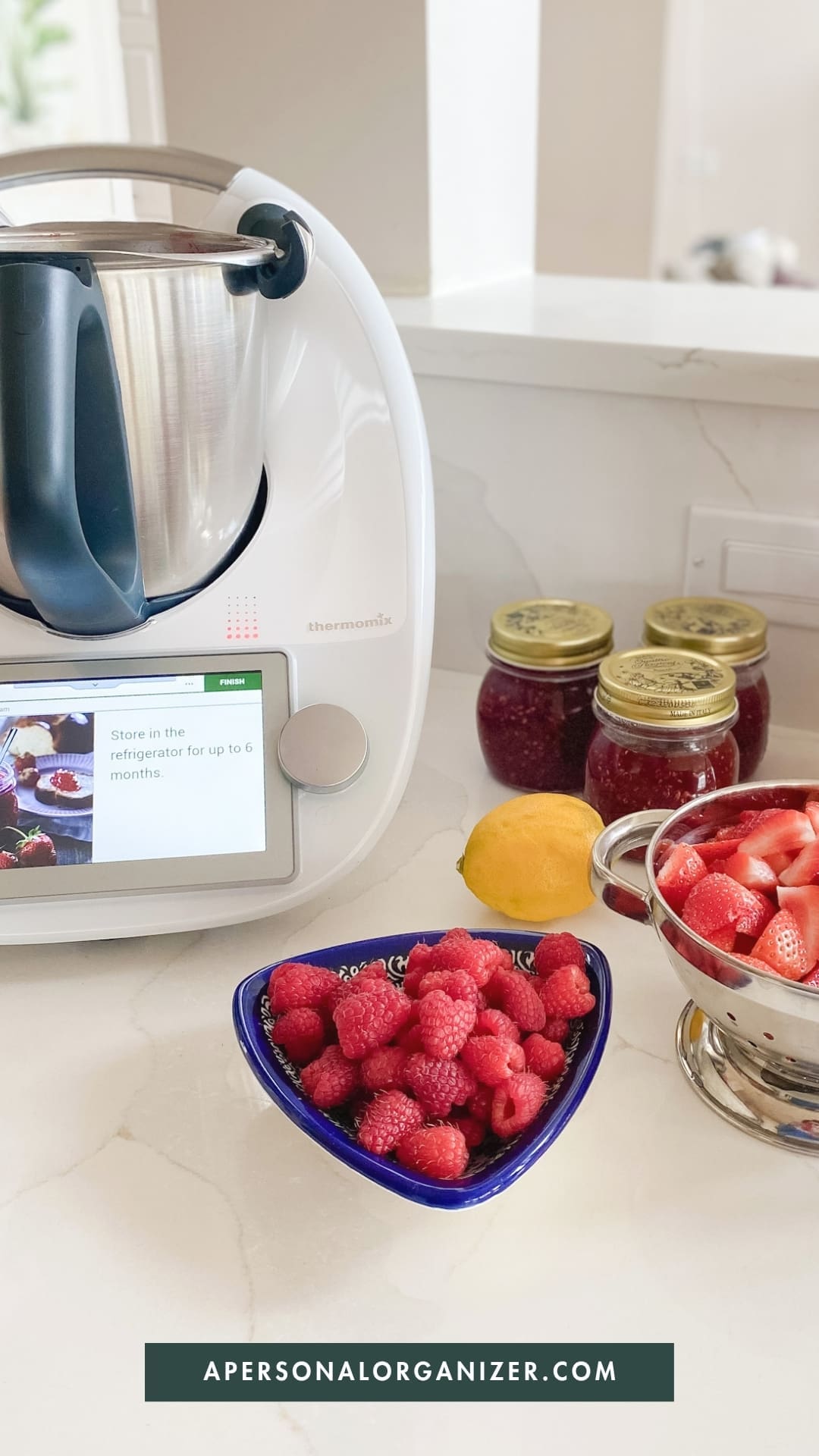 A Thermomix kitchen appliance sits on a counter near jars of jam, a lemon, fresh raspberries, and sliced strawberries—perfect for moms seeking time saving hacks from apersonalorganizer.com.